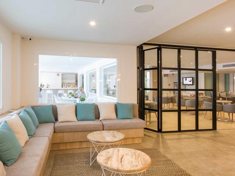 Sala luminosa del hotel con sofá en esquina, cojines verdes y mesas redondas de mármol junto a pared de vidrio.