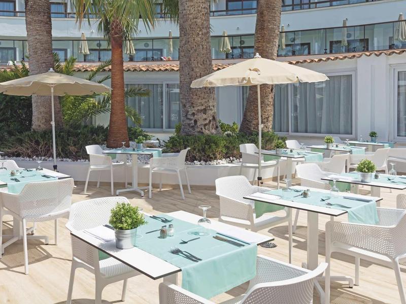 Terraza exterior iluminada de un hotel con mesas y sillas blancas, sombrillas y decoraciones.