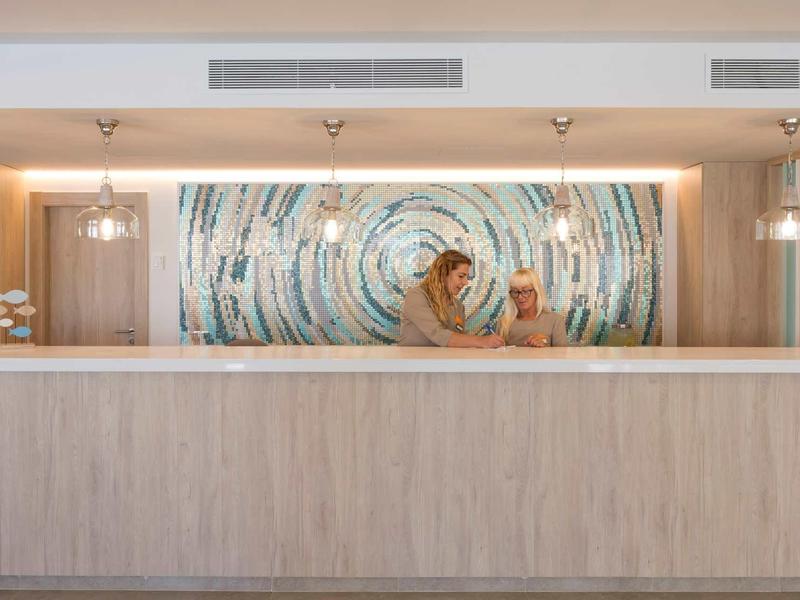 Recepción de hotel moderna con mostrador de madera clara y pared de mosaico abstracto al fondo.