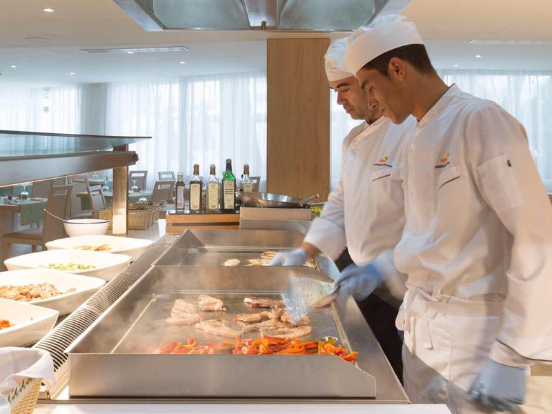 Dos chefs cocinando alimentos en una plancha grande en una cocina de restaurante luminosa.