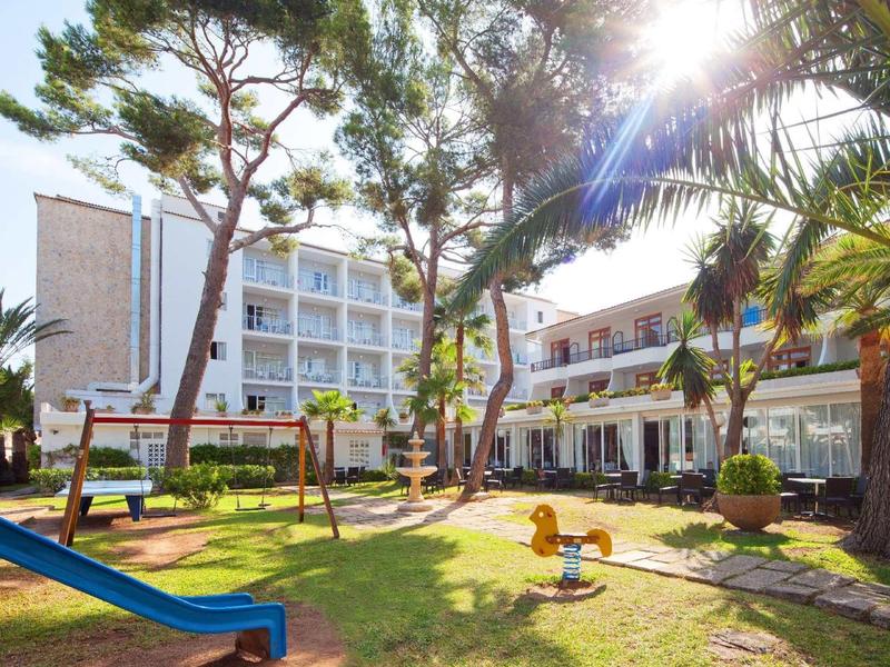 Hotelgebäude mit Terrasse, umgeben von Bäumen, Spielplatz mit Rutsche und Wippe bei Sonnenschein.