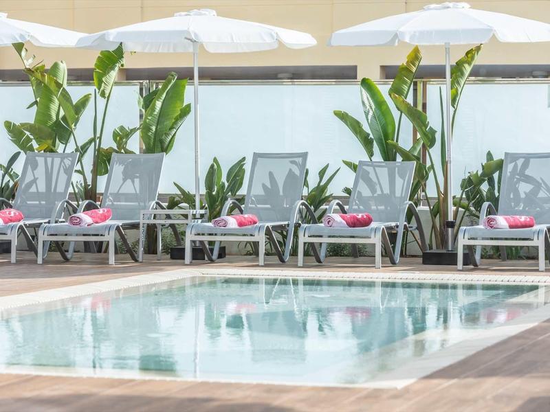 Schwimmbad mit Liegestühlen, weißen Sonnenschirmen und tropischen Pflanzen im Hintergrund.