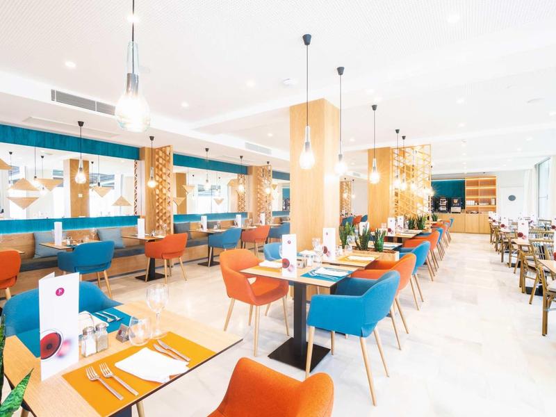 Helle, moderne Gastronomie mit bunten Stühlen, weißen Tischen und großer Fensterfront rechts.