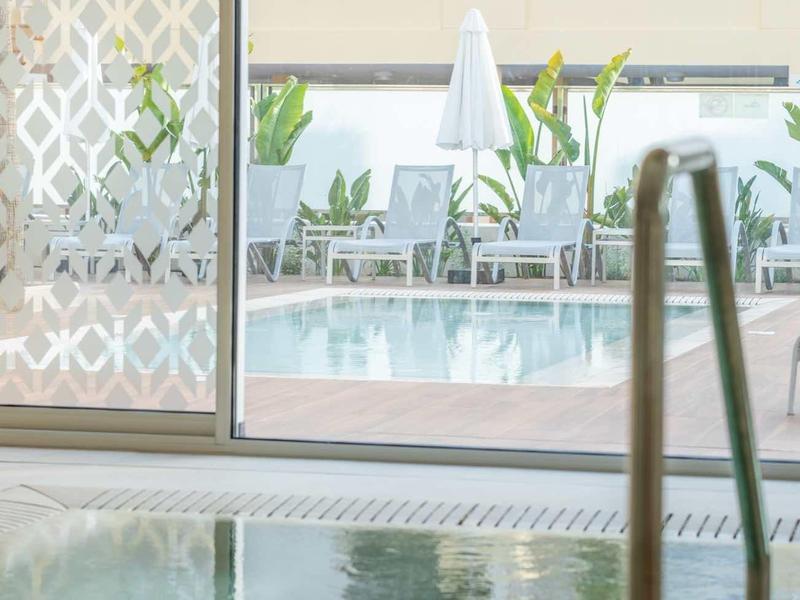 Blick durch Glastür auf Außenpool mit Liegestühlen, Sonnenschirm und Palmen im Hintergrund.