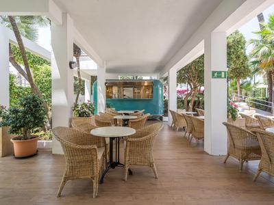 Überdachte Terrasse mit Korbstühlen und Tischen, umgeben von grünen Pflanzen und Palmen.