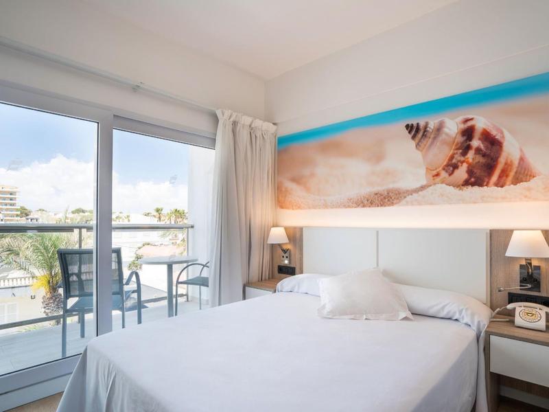 Helles Schlafzimmer mit Doppelbett, Balkon, weißer Einrichtung und großem Strandbild an der Wand.