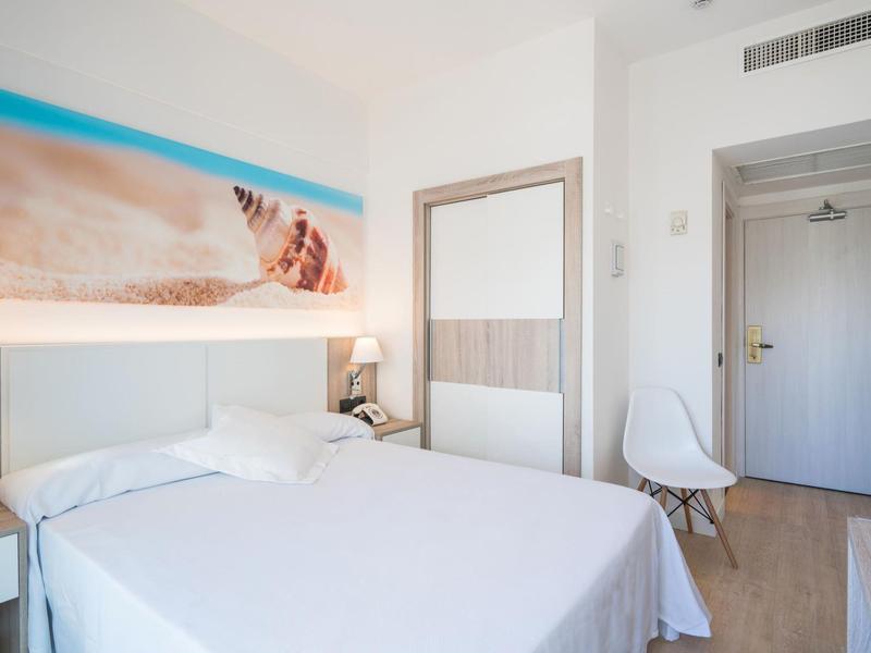 Schlichte, helle Hotelzimmer mit Doppelbett, weißem Stuhl und Strandbild an der Wand.