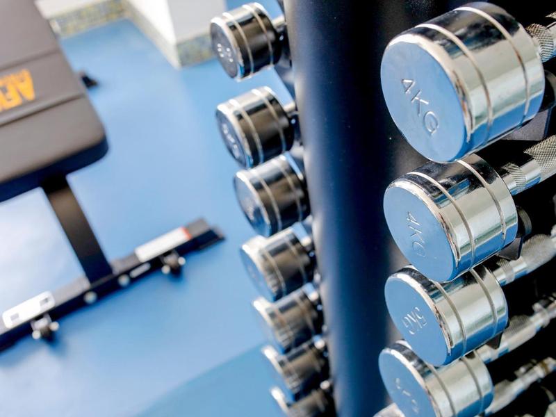 Kurzhanteln in glänzendem Chrom stehen aufgereiht neben einer schwarzen Hantelbank auf blauem Boden.