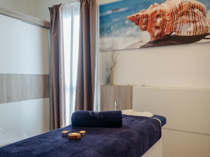 Behandlungsraum mit Massageliege, blauer Decke, Muschelbild und Vorhängen an Fenster.