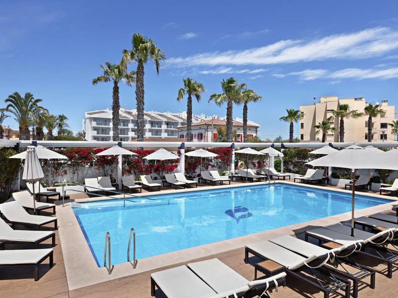 Rechteckiger Pool mit Liegen und Sonnenschirmen, Palmen und Hotelgebäuden im Hintergrund.