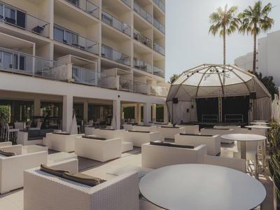 Área exterior moderna con muebles lounge y palmeras frente a un edificio hotelero.