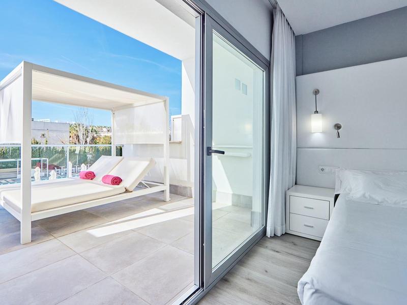 Balcón de hotel luminoso con cama de día y vista a piscina exterior, junto a habitación moderna con decoración blanca.