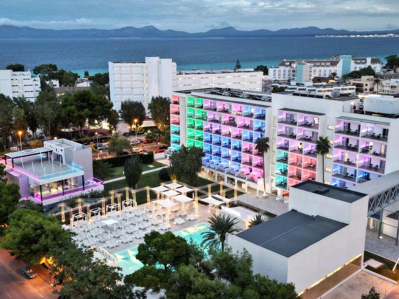 Gran hotel con balcones iluminados, rodeado de árboles y mar al fondo al atardecer.