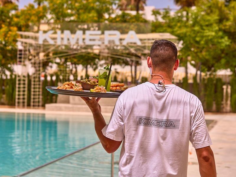 Hombre lleva una bandeja con bebidas y comidas junto a una piscina en un hotel.
