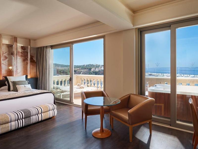 Bright hotel room with large bed, seating area, and balcony overlooking the sea.