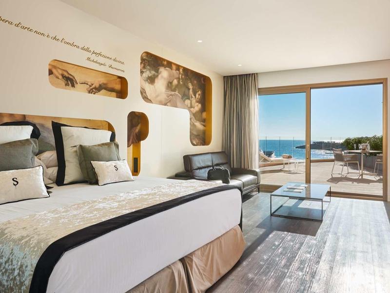 Modern hotel room with double bed, seating area, and terrace overlooking the sea.