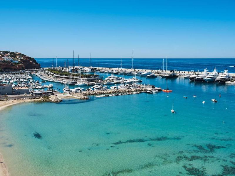 Clear blue sea with a harbor full of yachts and a small sandy beach.