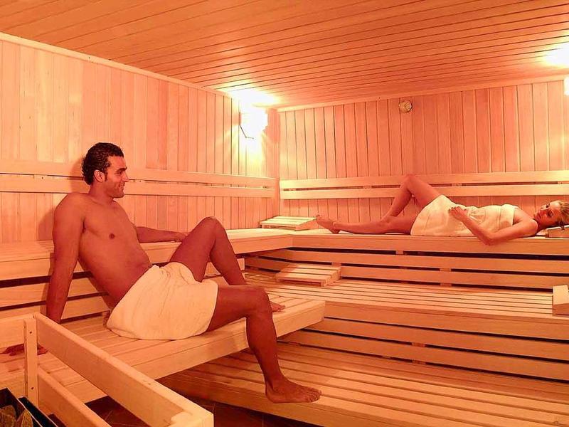 Two men relax in a wooden sauna on multi-level benches under warm lighting.
