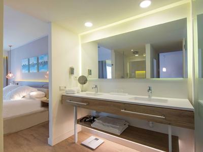 Modern bathroom with large mirror and sink, adjacent to a bedroom with double bed.