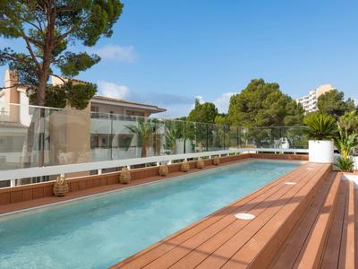 Modern hotel pool with wooden deck and glass barrier, surrounded by trees and buildings.