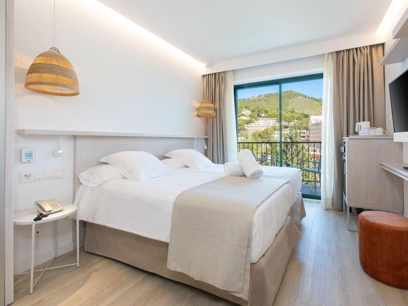 Bright hotel room with large bed, balcony, and mountain view.