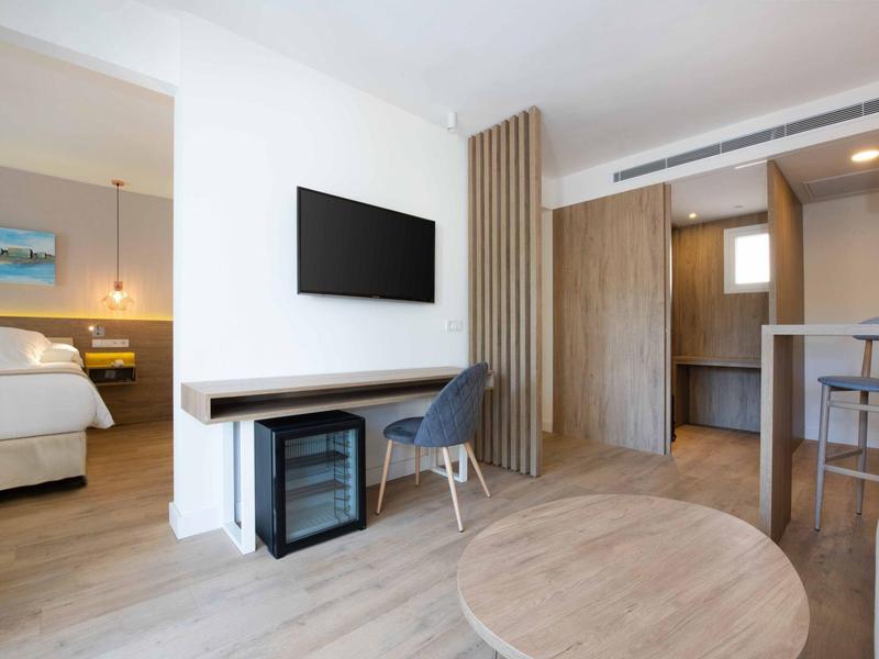 Bright hotel room with wooden flooring, modern TV, desk, and adjoining bedroom.