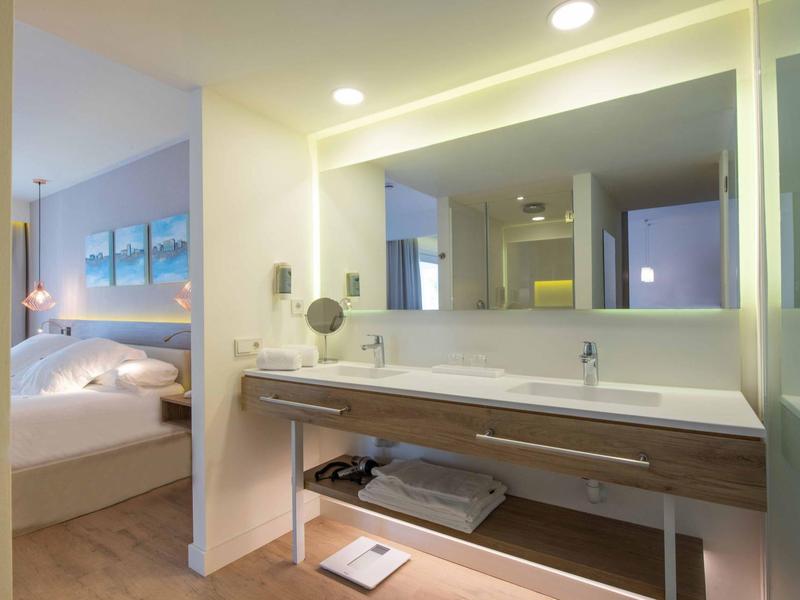 Modern bathroom with large mirror and sink, adjacent to a bedroom with double bed.