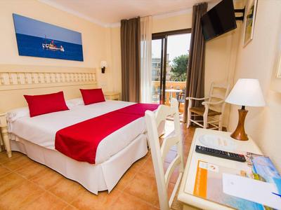 Jasny pokój hotelowy z podwójnym łóżkiem, czerwoną narzutą, biurkiem i balkonem z widokiem na zewnątrz.