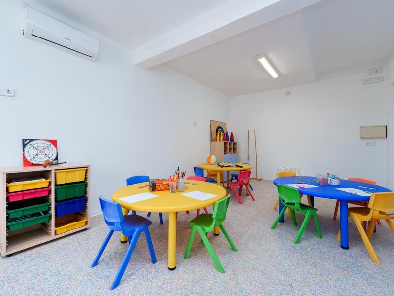 Buntes Kinderspielzimmer mit kleinen Tischen, Stühlen und Spielregalen in hellem Raum.