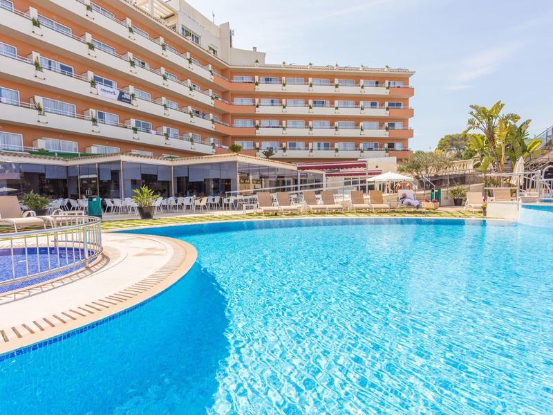 Großer Außenpool vor einem mehrstöckigen Hotel mit Sonnenliegen und Palmen.