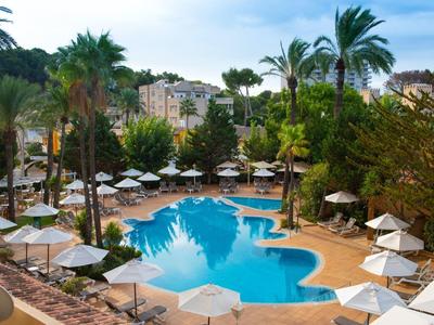 Schwimmbad mit unregelmäßiger Form, umgeben von Palmen, Sonnenschirmen und Gebäuden im Hintergrund.