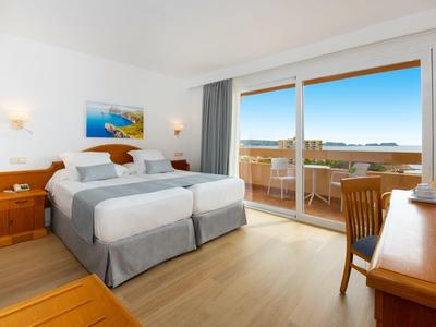 Helles Hotelzimmer mit zwei Betten, Schreibtisch und Balkon mit Meerblick.