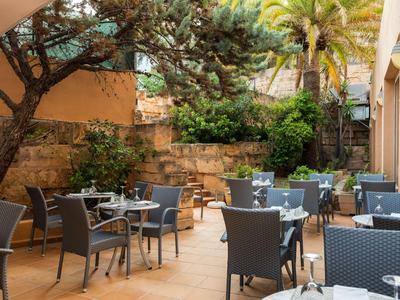 Außenbereich eines Cafés mit grau-gelben Tischen und Stühlen unter Bäumen und Palmen.