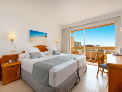 Helles Hotelzimmer mit zwei Einzelbetten, Balkon und Meerblick bei Sonnenschein.