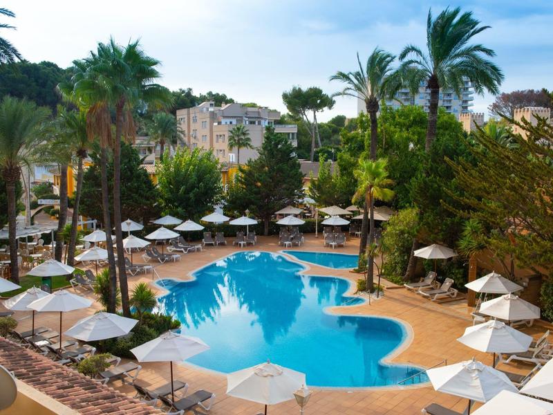 Schwimmbad mit unregelmäßiger Form, umgeben von Palmen, Sonnenschirmen und Gebäuden im Hintergrund.