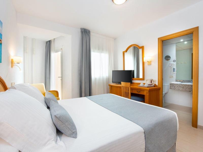 Helles Hotelzimmer mit Doppelbett, grauen Vorhängen, Fernseher und angrenzendem Badezimmer.