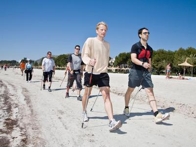 Gruppo di persone che camminano sulla spiaggia con bastoncini da nordic walking sotto un cielo sereno.