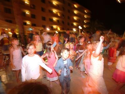 Niños bailan al aire libre por la noche frente a un hotel iluminado.
