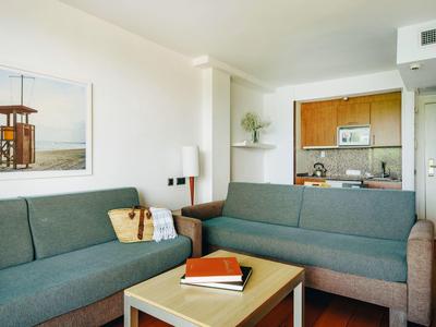 Acogedora área de estar en hotel con dos sofás, mesa de centro, cocina pequeña y cuadro en pared clara.