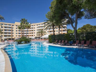 Gran piscina de hotel frente a un edificio de hotel de varias plantas con palmeras y tumbonas bajo un cielo claro.