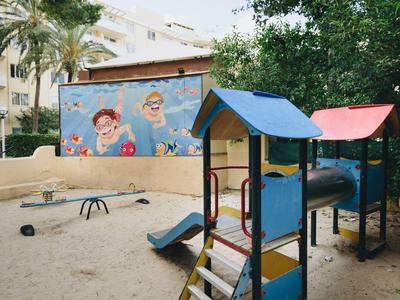 Área de juegos infantiles con tobogán y estructuras para escalar, rodeada de arena y edificios residenciales.