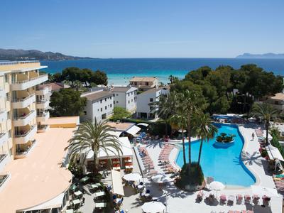Vista de un hotel con piscina, palmeras y mar al fondo bajo un cielo despejado.