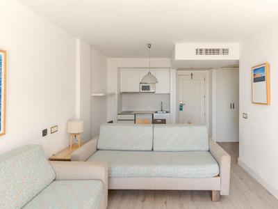 Área de habitación de hotel moderna con mobiliario claro, sofá y cocina integrada