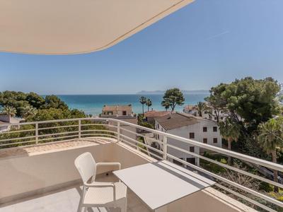 Balcón con silla y mesa con vista a árboles mediterráneos, casas y el mar.