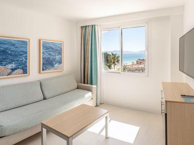Habitación de hotel luminosa con sofá, mesa de café, cuadros y vista desde la ventana de palmeras y mar.