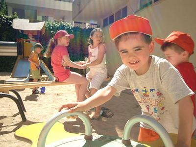 Niños jugando en un parque soleado frente a un edificio del hotel, algunos llevan gorras rojas.