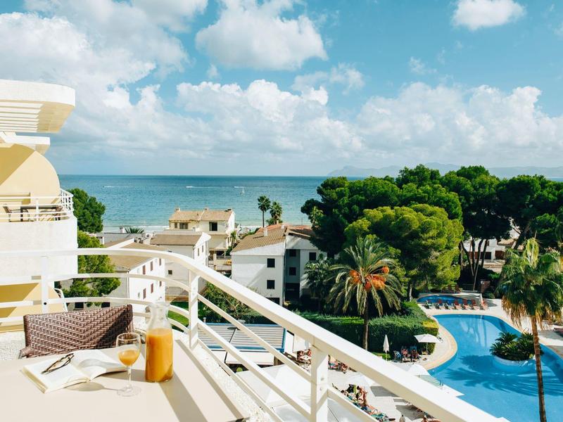 Balcón con desayuno y vista a la piscina, palmeras y el mar bajo un cielo con nubes.