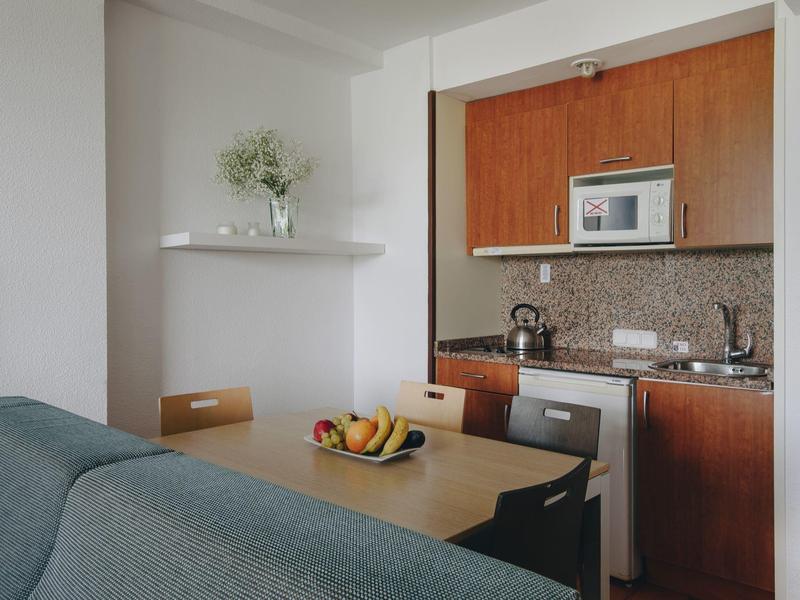 Pequeña cocina moderna con armarios de madera, mesa de comedor y cesta de frutas en un apartamento de hotel.