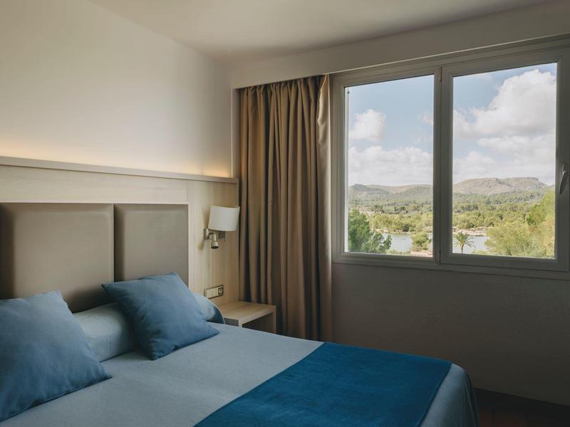 Dormitorio con cama doble y tres almohadas azules junto a una ventana con vista al paisaje.