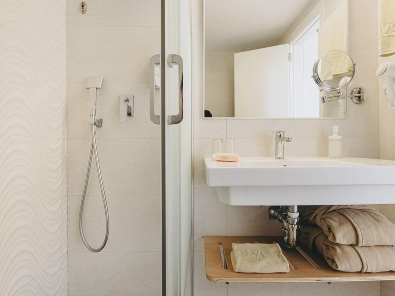 Baño luminoso con ducha de vidrio, lavabo blanco y toallas en estante de madera.